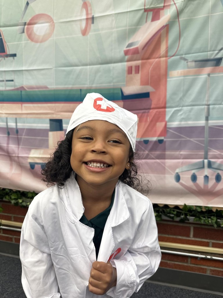 A smiling TK student dressed in a doctor costume during imaginative play